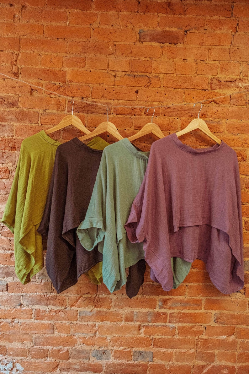 May include: Five different colored cotton tops hanging on wooden hangers against a brick wall. The tops are in shades of green, brown, purple, and a darker green.