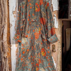 May include: A long, patterned coat in shades of gray and green, adorned with vibrant orange and yellow floral designs. The coat features a button closure, pockets, and rolled-up sleeves. The garment is hanging on a wooden hanger.