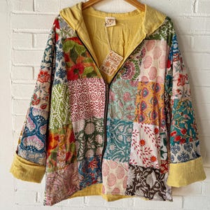 May include: A patchwork zip-up hoodie with a yellow hood and cuffs. The jacket features a variety of floral and geometric patterns in shades of red, green, blue, and cream. The jacket is hanging on a wooden hanger against a white brick wall.