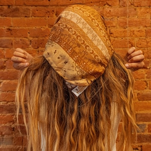 May include: A person with long, wavy, light brown hair wearing a brown and white patterned bandana with a floral design. The bandana has a small tag that says "Handmade". The person is wearing a white shirt and blue jeans.