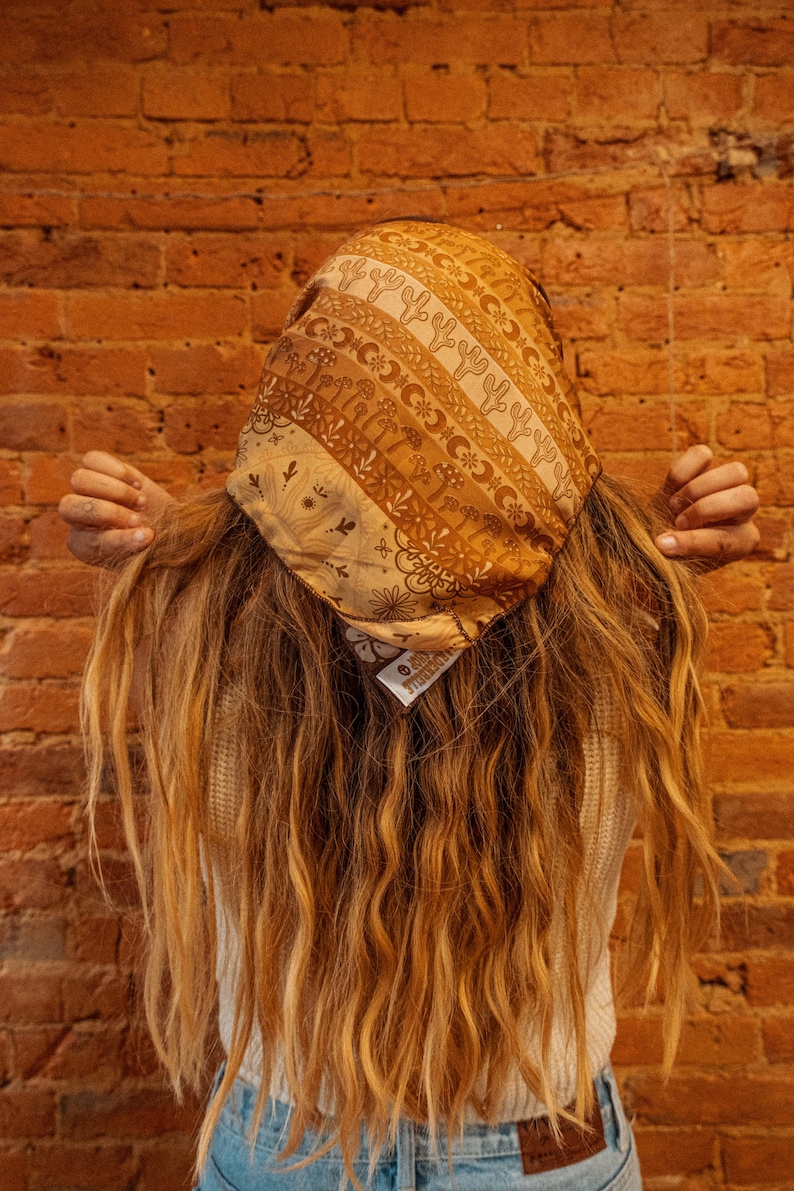 May include: A person with long, wavy, light brown hair wearing a brown and white patterned bandana with a floral design. The bandana has a small tag that says "Handmade". The person is wearing a white shirt and blue jeans.