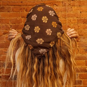 May include: A person with long, wavy, light brown hair is wearing a brown bandana with a floral pattern. The bandana has yellow and pink flowers on a dark brown background. The person is wearing a white sweater and blue jeans.