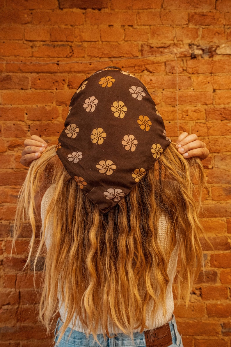 May include: A person with long, wavy, light brown hair is wearing a brown bandana with a floral pattern. The bandana has yellow and pink flowers on a dark brown background. The person is wearing a white sweater and blue jeans.