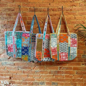 May include: Four colorful patchwork tote bags hanging on hooks against a brick wall. The bags are made of different fabrics and have a variety of patterns, including floral, geometric, and paisley.