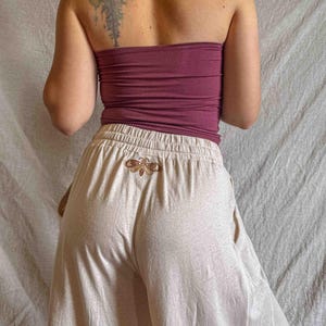 K&ouml;nnte beinhalten: Ein weinrotes tr&auml;gerloses Schlauchoberteil kombiniert mit einer hellbeigen weiten Hose mit elastischem Bund und einem kleinen gestickten Mottenmotiv, das mittig direkt unter der Taille auf der R&uuml;ckseite der Hose platziert ist.