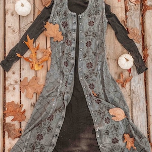 May include: A long, gray, button-up dress with a floral pattern. The dress is open in the front, revealing a black turtleneck underneath. The dress is laid out on a wooden surface with fall leaves and white pumpkins.