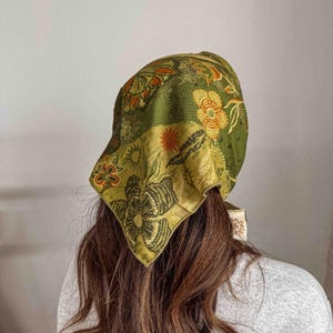 Puede incluir: Un pa&ntilde;uelo con estampado floral verde y amarillo. El cubrecabezas presenta una mezcla de dise&ntilde;os florales y geom&eacute;tricos en tonos verdes, amarillos y naranjas. El pa&ntilde;uelo est&aacute; atado en la parte posterior de la cabeza, con los extremos del pa&ntilde;uelo visibles.