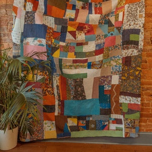 Puede incluir: Una gran colcha de retazos hecha de varias telas en tonos de azul, rojo, verde, marrón y amarillo. La colcha está colgada en una pared de ladrillo y tiene una variedad de patrones, incluyendo florales, geométricos y a cuadros.