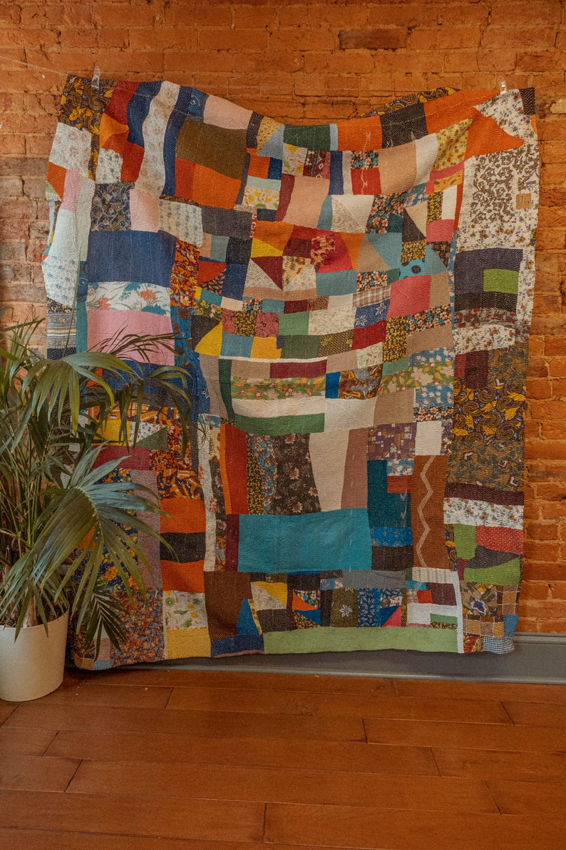 May include: A large patchwork quilt made of various fabrics in shades of blue, red, green, brown, and yellow. The quilt is hanging on a brick wall and has a variety of patterns, including floral, geometric, and plaid.