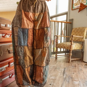 May include: A person wearing a white crop top and brown patchwork pants with a drawstring waist. The pants are made of different fabrics and colors, including brown, blue, and red. The person is standing on a wooden floor.