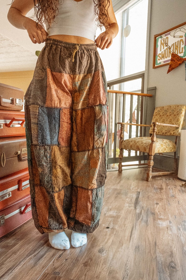 May include: A person wearing a white crop top and brown patchwork pants with a drawstring waist. The pants are made of different fabrics and colors, including brown, blue, and red. The person is standing on a wooden floor.