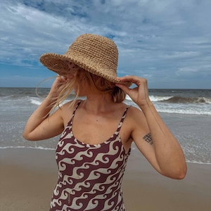Pu&ograve; includere: Una persona sulla spiaggia indossa un cappello di paglia intrecciata e un costume da bagno marrone e bianco a fantasia di onde. La persona tiene in mano il cappello. Il mare e il cielo sono visibili sullo sfondo.