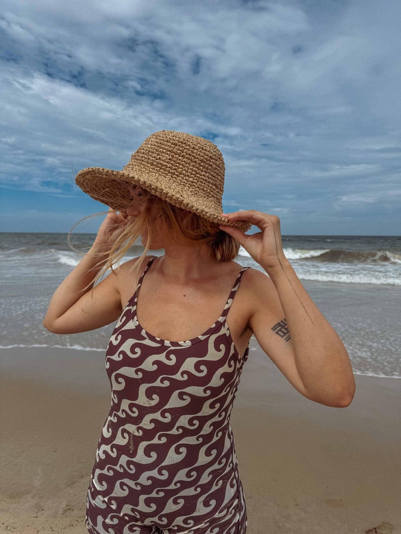 Pu&ograve; includere: Una persona sulla spiaggia indossa un cappello di paglia intrecciata e un costume da bagno marrone e bianco a fantasia di onde. La persona tiene in mano il cappello. Il mare e il cielo sono visibili sullo sfondo.
