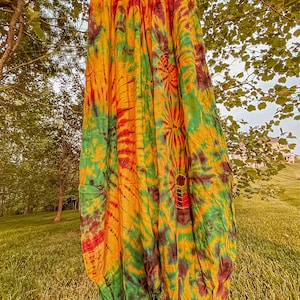 May include: A pair of tie-dye trousers in yellow, green, and red hangs from a tree branch. The loose-fitting trousers feature a gathered waist and a relaxed design. The vibrant colours create a unique pattern, ideal for casual wear. The trousers are made of a light fabric.