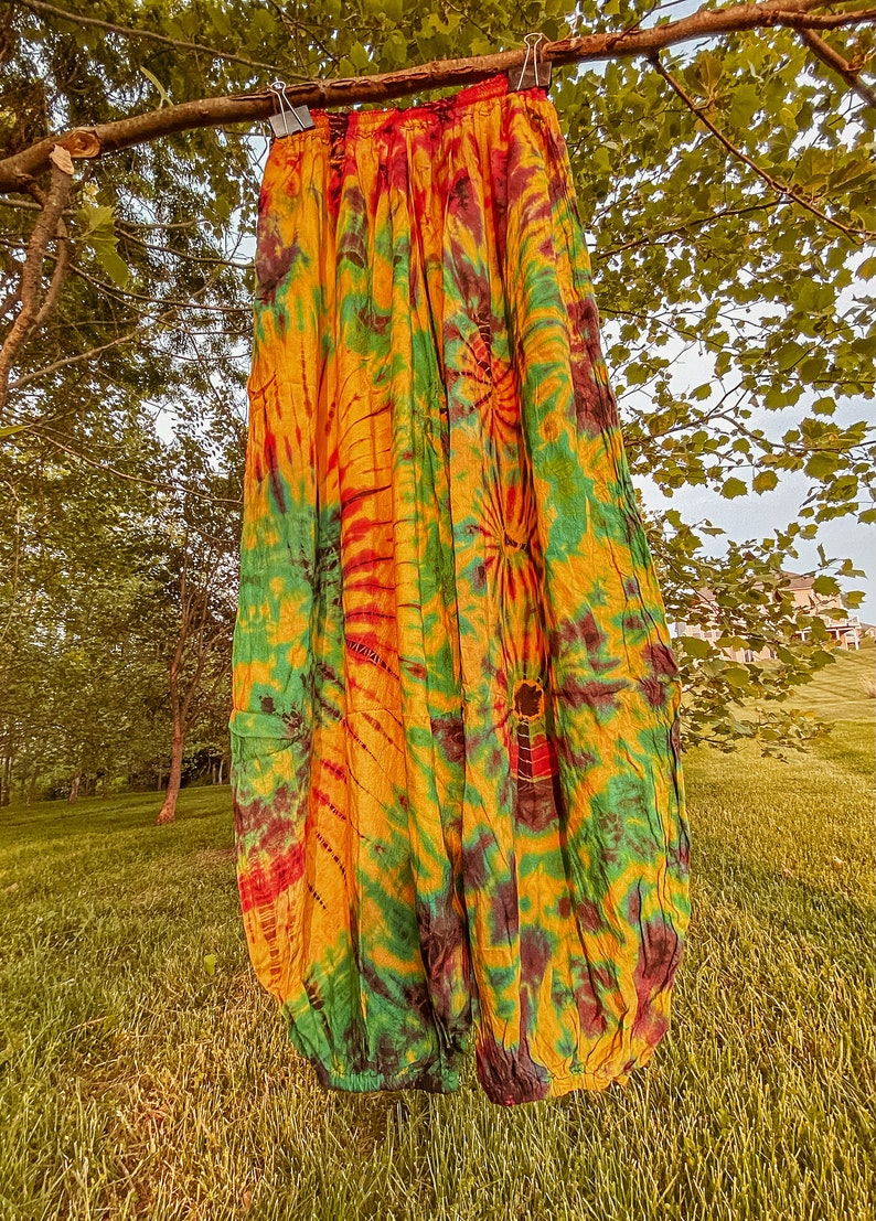 May include: A pair of tie-dye trousers in yellow, green, and red hangs from a tree branch. The loose-fitting trousers feature a gathered waist and a relaxed design. The vibrant colours create a unique pattern, ideal for casual wear. The trousers are made of a light fabric.