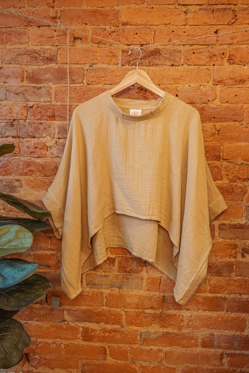 May include: A light brown, loose-fitting, short-sleeved top with a slightly uneven hemline. The top is hanging on a wooden hanger against a brick wall.