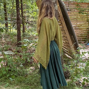 May include: A long-sleeved olive green top with wide, flared sleeves featuring a delicate brown geometric pattern along the cuffs, paired with a floor-length, solid dark teal skirt that has a matching brown geometric border at the hemline.