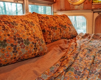 May include: A bed with a brown floral patterned duvet cover and two matching pillowcases. The bed is in a converted bus with wood paneling and string lights.