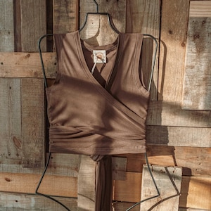 May include: A brown sleeveless wrap top with a V-neckline and a tie at the waist. The top is hanging on a black hanger against a wooden background.