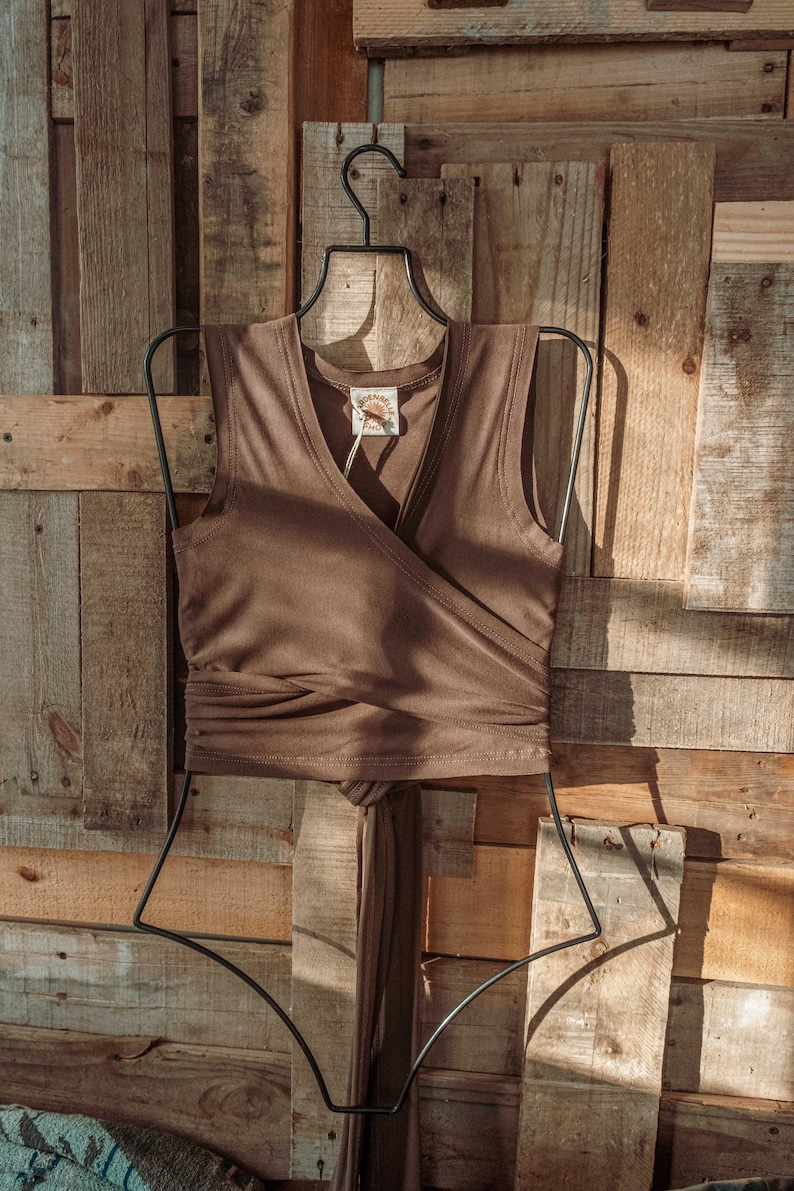 May include: A brown sleeveless wrap top with a V-neckline and a tie at the waist. The top is hanging on a black hanger against a wooden background.