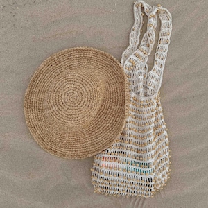Pu&ograve; includere: Un cappello da sole in paglia intrecciata e una borsa tote all'uncinetto bianca e beige con perline di legno sono appoggiati sulla sabbia. Il cappello &egrave; rotondo con una tesa larga. La borsa ha manici lunghi.