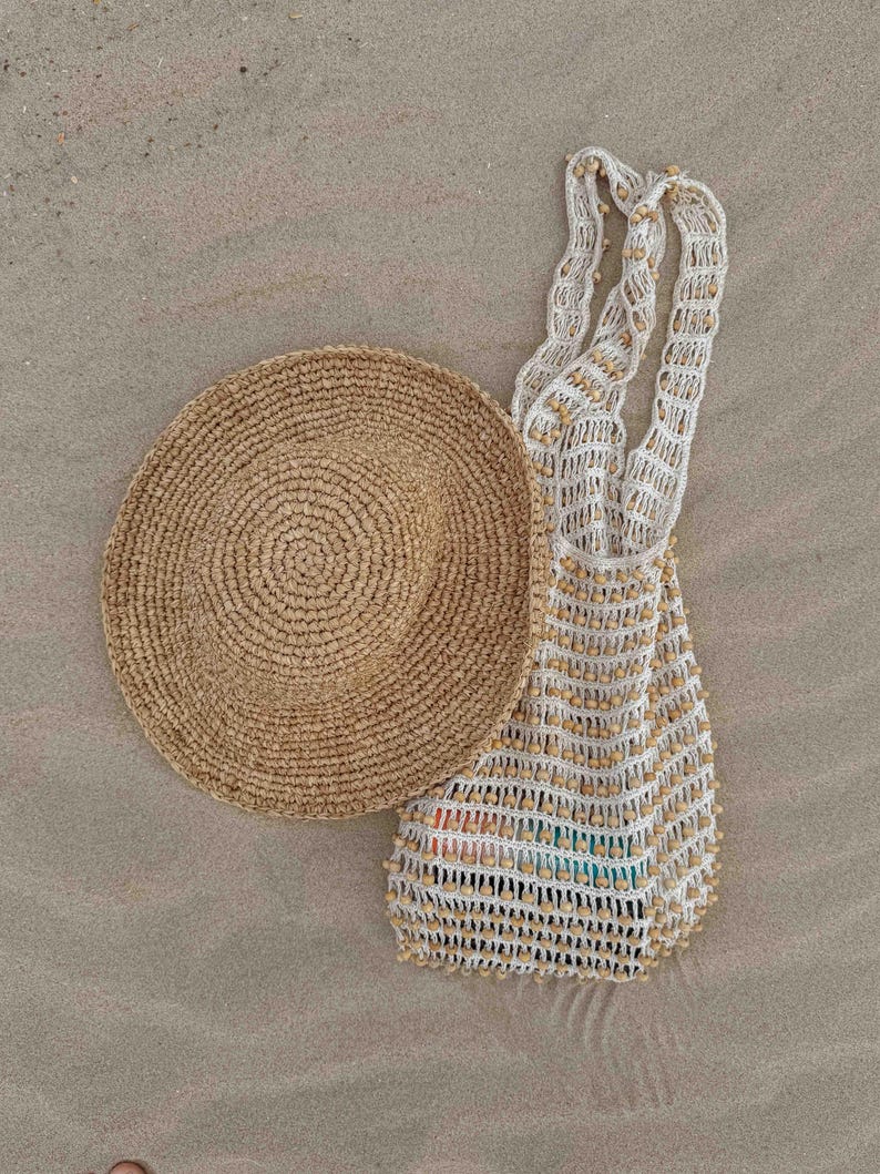 Pu&ograve; includere: Un cappello da sole in paglia intrecciata e una borsa tote all'uncinetto bianca e beige con perline di legno sono appoggiati sulla sabbia. Il cappello &egrave; rotondo con una tesa larga. La borsa ha manici lunghi.
