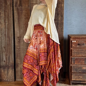 May include: A cream-colored, long-sleeved sweater paired with a flowing, asymmetrical skirt. The skirt features a vibrant floral pattern in shades of pink, orange, and burgundy. The outfit is completed with brown boots, creating a bohemian aesthetic.