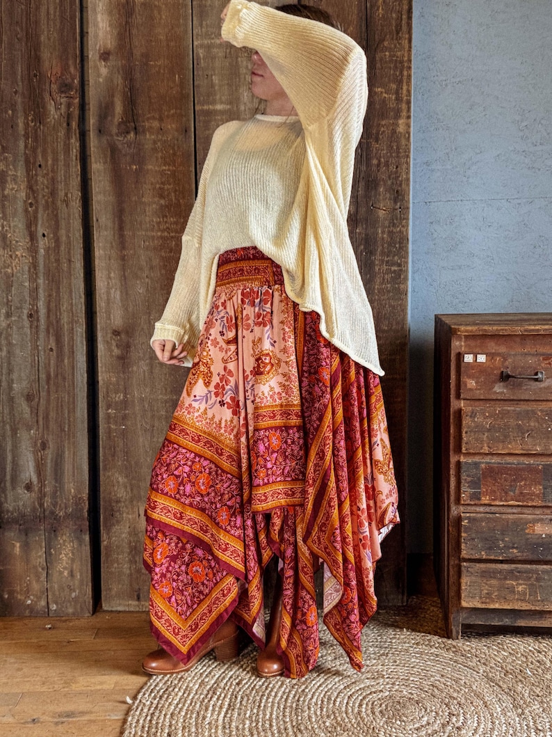 May include: A cream-colored, long-sleeved sweater paired with a flowing, asymmetrical skirt. The skirt features a vibrant floral pattern in shades of pink, orange, and burgundy. The outfit is completed with brown boots, creating a bohemian aesthetic.