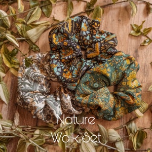 May include: Set of three floral patterned hair scrunchies in shades of green, brown, and cream. The scrunchies are arranged on a wooden surface with green foliage.