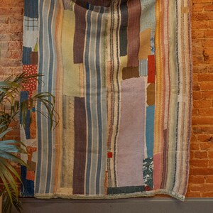 K&ouml;nnte beinhalten: Ein bunter Patchwork-Deckenbezug mit verschiedenen Streifen und Mustern in Blaut&ouml;nen, Braun, Beige und Rosa. Der Deckenbezug h&auml;ngt an einer Backsteinwand.