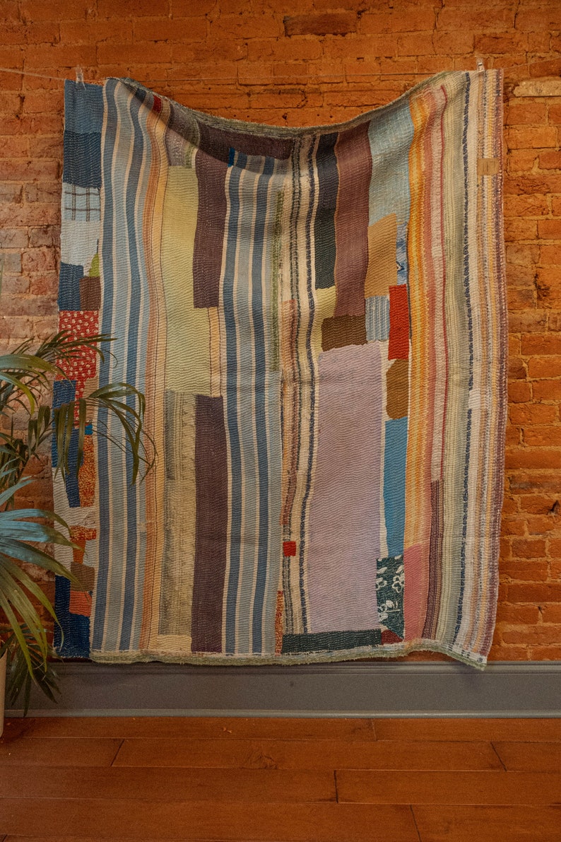 K&ouml;nnte beinhalten: Ein bunter Patchwork-Deckenbezug mit verschiedenen Streifen und Mustern in Blaut&ouml;nen, Braun, Beige und Rosa. Der Deckenbezug h&auml;ngt an einer Backsteinwand.