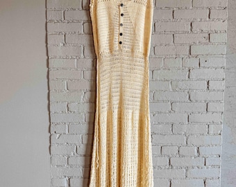 Könnte beinhalten: Ein cremefarbenes, ärmelloses Häkelkleid mit rundem Ausschnitt und einer Reihe schwarzer Knöpfe am Oberteil. Das Kleid hängt an einem Holzbügel vor einer grauen Backsteinmauer. Das Kleid hat eine taillierte Taille und einen ausgestellten Rock.