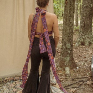 May include: A woman wearing a purple and orange floral halter top with a long tie that wraps around the neck and ties in the back. The top is worn with black flared pants.