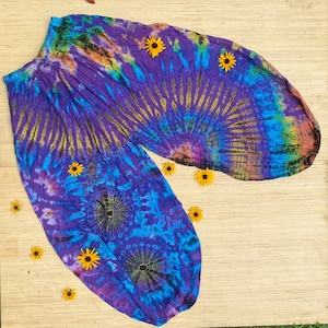 May include: Tie-dye harem trousers in purple, blue, yellow, and green. The trousers have an elasticated waistband and gathered ankles. Yellow flowers are scattered around on a light brown surface.