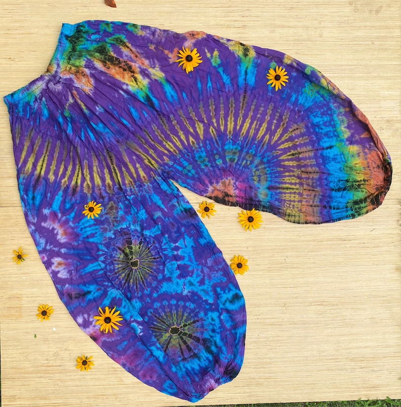 May include: Tie-dye harem trousers in purple, blue, yellow, and green. The trousers have an elasticated waistband and gathered ankles. Yellow flowers are scattered around on a light brown surface.