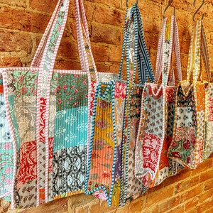 May include: Five colorful patchwork tote bags hang on hooks against a brick wall. The bags are made of different fabrics and patterns, including floral, geometric, and paisley. Each bag has a unique design and color combination.