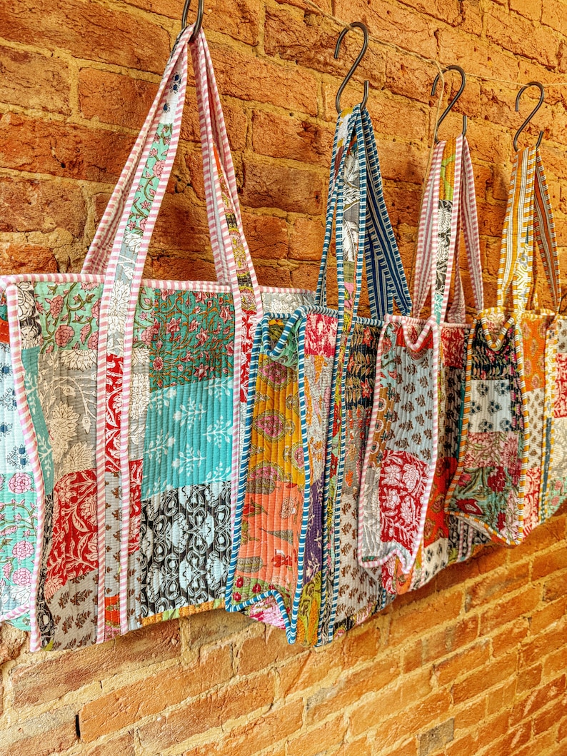 May include: Five colorful patchwork tote bags hang on hooks against a brick wall. The bags are made of different fabrics and patterns, including floral, geometric, and paisley. Each bag has a unique design and color combination.