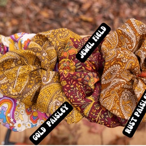 May include: Five fabric scrunchies with different patterns. The scrunchies are in a row from left to right: "ZOEY", "GOLD PAISLEY", "JEWEL FIELD", "RUST PAISLEY", and "BUTTER FLOWER".