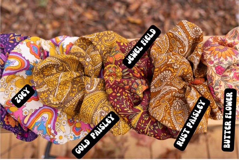 May include: Five fabric scrunchies with different patterns. The scrunchies are in a row from left to right: "ZOEY", "GOLD PAISLEY", "JEWEL FIELD", "RUST PAISLEY", and "BUTTER FLOWER".