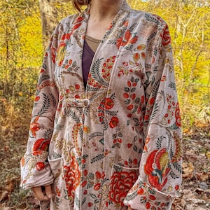 Puede incluir: Una bata larga de color claro con un estampado floral en rojo, naranja y verde. La bata tiene cierre de botón y bolsillos. Las mangas están dobladas y la tela parece suave y fluida. La bata se lleva sobre una parte superior oscura.