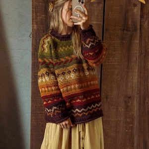 May include: A colorful, patterned sweater with a high neckline. The sweater features a mix of green, orange, and burgundy hues. The person is wearing a long, tan skirt. A wooden hanger is visible in the background.