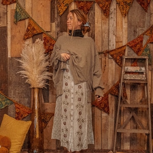 May include: A woman wearing a brown sweater and a white floral skirt stands in front of a wooden wall decorated with colorful fabric pennants.