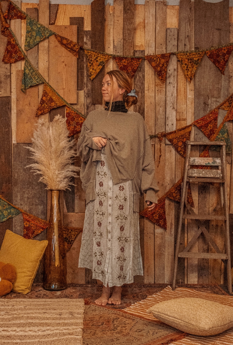 May include: A woman wearing a brown sweater and a white floral skirt stands in front of a wooden wall decorated with colorful fabric pennants.