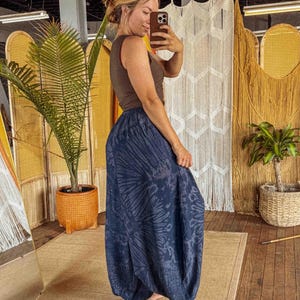 May include: A woman wearing a brown vest top and navy blue harem trousers with a tie-dye pattern. The trousers are loose-fitting and gathered at the ankles. The background includes a mirror, a woven screen, and a potted plant.