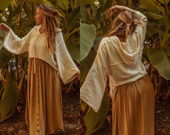 May include: A woman wearing a white, loose-fitting, hooded top with long sleeves and a brown, long, button-down skirt. She is standing in a tropical garden with lush green foliage.