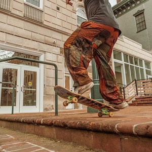 Op de afbeelding: Een skateboarder in een broek met patroon voert een truc uit op een skateboard met groene wielen. De scène speelt zich af op een bakstenen oppervlak met een gebouw op de achtergrond.