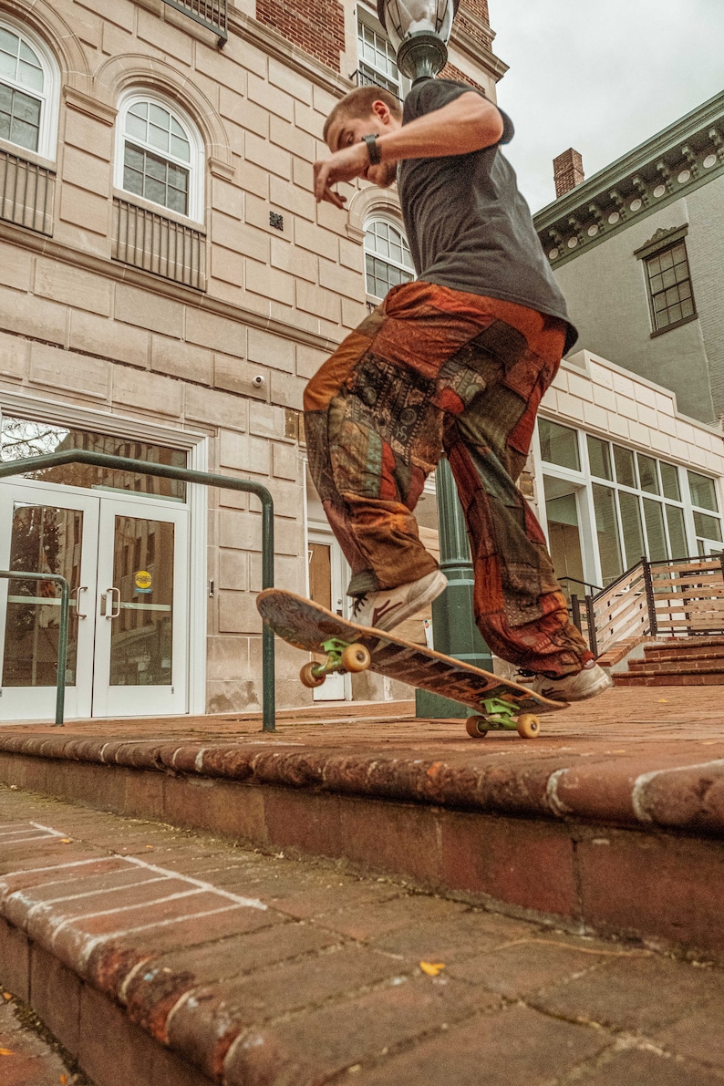 May include: A person wearing a black shirt and colorful patchwork pants is skateboarding down a set of brick stairs. The skateboard is brown with green wheels.