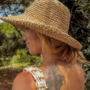 Pu&ograve; includere: Cappello di paglia intrecciata color beige naturale con tesa larga. La trama del cappello &egrave; visibile. La persona che indossa il cappello ha i capelli biondi. Indossa un top in macram&egrave; bianco con perline di legno. Lo sfondo &egrave; verde sfocato.