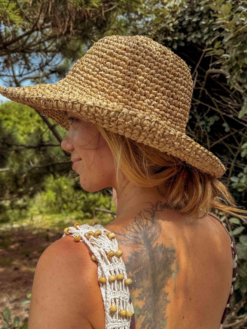 Pu&ograve; includere: Cappello di paglia intrecciata color beige naturale con tesa larga. La trama del cappello &egrave; visibile. La persona che indossa il cappello ha i capelli biondi. Indossa un top in macram&egrave; bianco con perline di legno. Lo sfondo &egrave; verde sfocato.