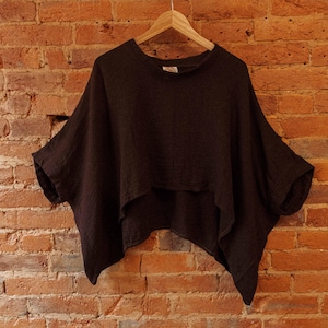 May include: A dark brown, loose-fitting, short-sleeved top with a rounded neckline. The top is hanging on a wooden hanger against a brick wall.